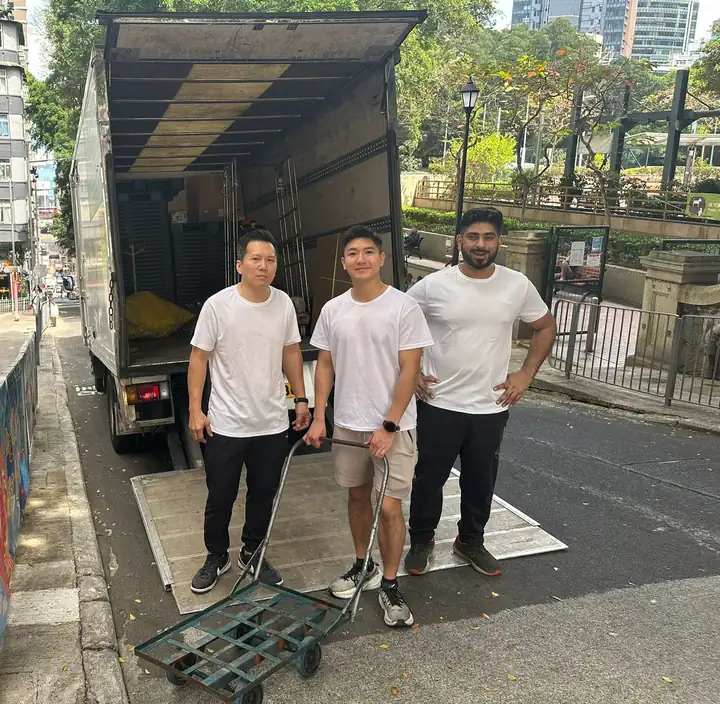 三位好哥哥搬屋團隊成員穿白色T恤，站喺香港街頭一架打開咗嘅搬屋貨車同手推車旁邊